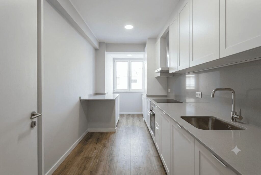 Visão ampla em perspectiva de uma cozinha corredor moderna e planejada. A bancada principal em quartzo cinza claro abriga a pia e o cooktop de forma linear, com armários brancos sob medida e piso de madeira que amplia o ambiente.
