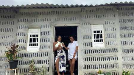 Uma mãe e sua filha, ambas vestidas de branco, sorrindo e segurando copos em comemoração na porta de sua casa com paredes feitas de garrafas de vidro recicladas.