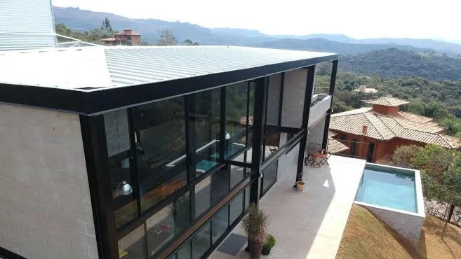 Vista aérea de uma casa galpão moderna com estrutura metálica preta e paredes de blocos de concreto, telhado de zinco e piscina na encosta.