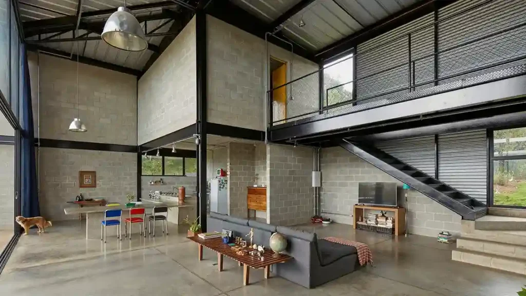 Interior de casa galpão com pé direito duplo, estrutura metálica aparente no teto, mezanino industrial e paredes de blocos de concreto.